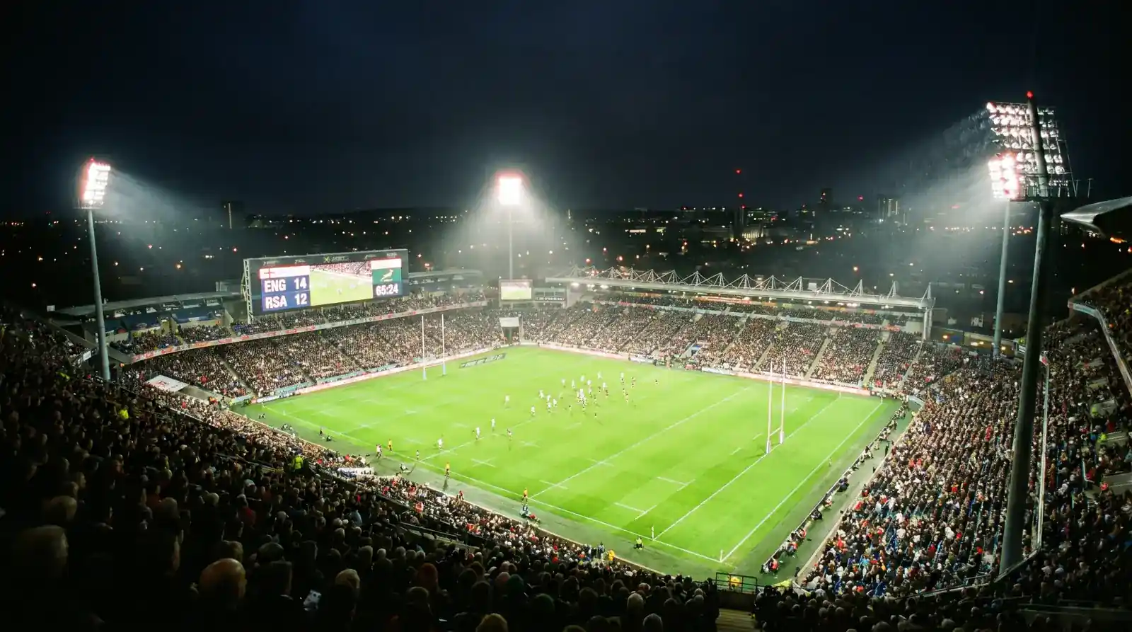 Floodlit rugby stadium at night with fans watching an in-play match and electronic scoreboard glowing